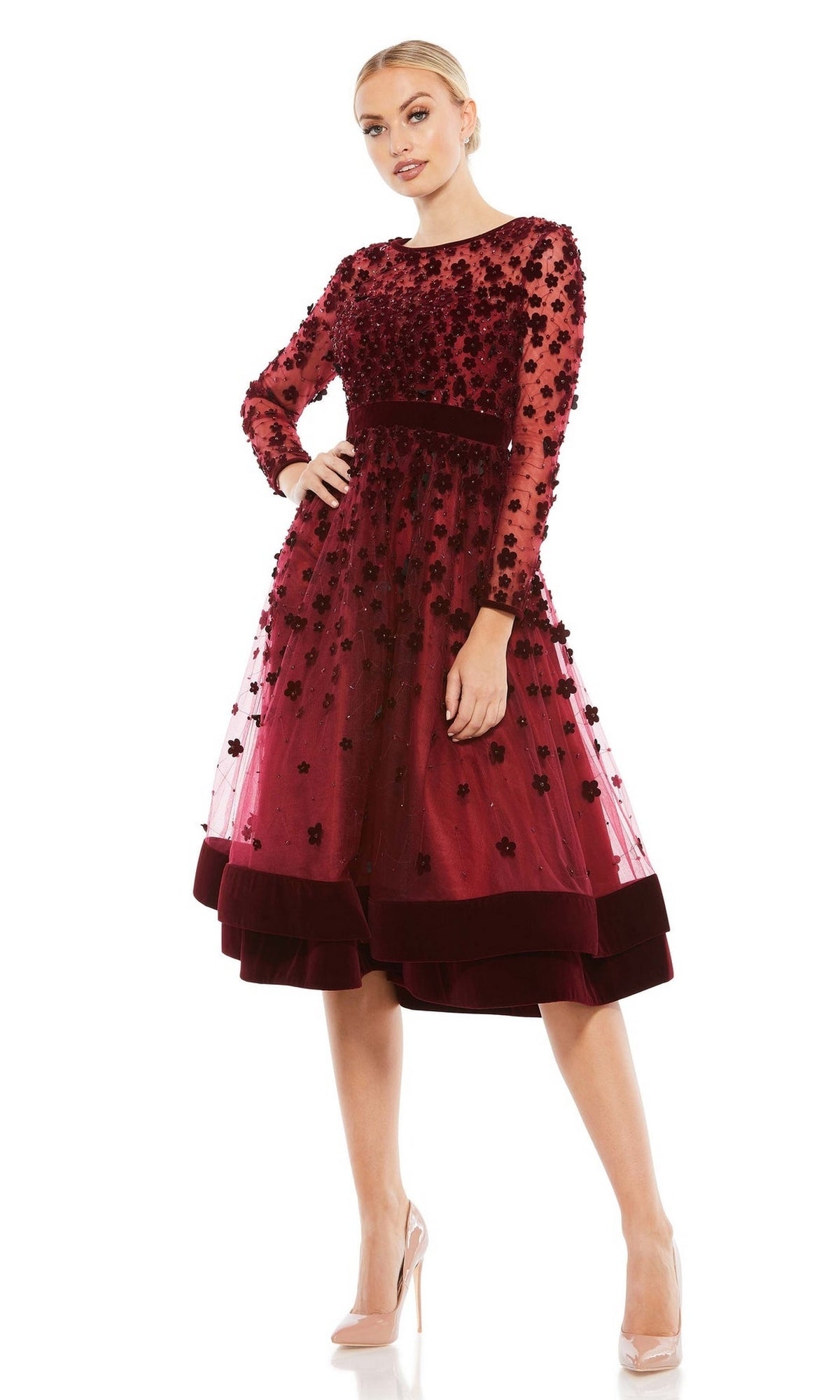 A woman stands against a white background in the Mac Duggal 67007 knee-length wedding-guest dress, featuring long sleeves, burgundy floral appliqués, and a sheer overlay, paired with nude heels.